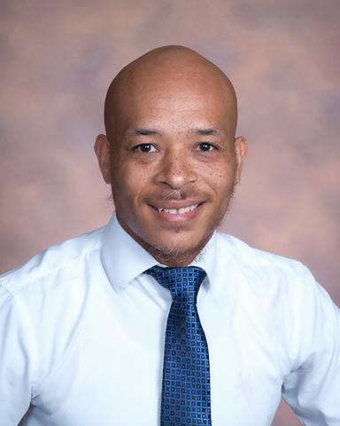 Darryl Hunt smiles in a professional headshot wearing a white dress shirt and blue patterned tie against a mottled brown studio background.