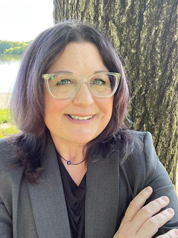 Christina Israel smiles in an outdoor portrait taken near a tree and river, wearing a gray blazer and clear-framed glasses.