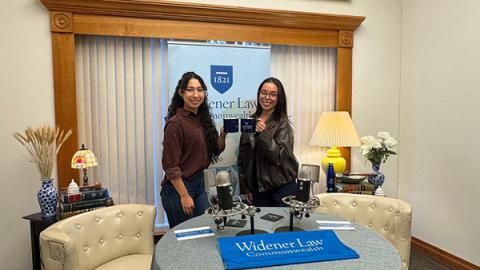 Michelle and Johana standing in the podcast room behind the microphone and desk with a Widener Law Commonwealth coffee mug. 
