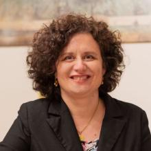 Maria Sabatino — Smiling woman with curly dark hair in a black blazer, photographed in front of abstract artwork.Maria Sabatino — Smiling woman with curly dark hair in a black blazer.