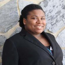 Waleisha Moat smiles in a portrait taken outdoors against a stone wall, wearing a black blazer.
