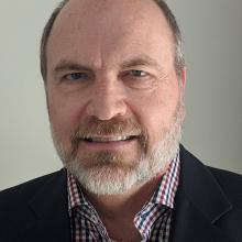 Thomas J. Foley — headshot in a dark blazer and plaid shirt, against a light gray background.