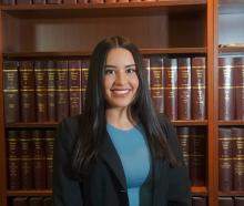 Emilie Baerga — smiling headshot in a black blazer over a blue top, posed in front of law library shelves.