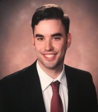 Ryan Sypniewski — Formal portrait of a young man in a black suit and red tie, against a warm studio background.