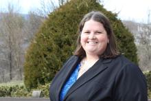 Leah Bobula smiling outdoors in a black blazer over a blue top, with campus grounds and bare winter trees in the background.