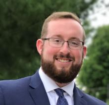 Philip Mintz — smiling headshot in a navy suit and blue tie, photographed outdoors with greenery in the background.