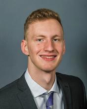 Thomas Dyer — Professional headshot of a young man in a gray suit and blue tie, against a gray studio background.