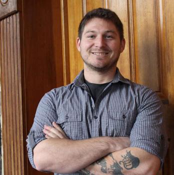 WLC alumnus Nick in front of a wooden door with his arms crossed smiling.