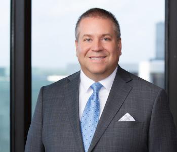 WLC alumnus in front of window with a city view in a gray suit with a blue tie.