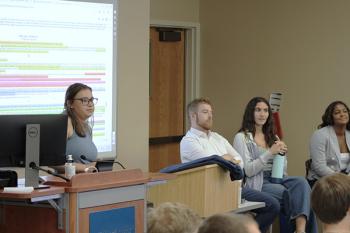 Three students at the front of the class in chairs and one student at the podium with a slideshow behind her