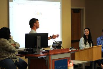 A student behind the podium gesturing with their hand and another student close by in a chair