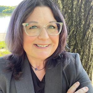 Christina Israel smiles in an outdoor portrait taken near a tree and river, wearing a gray blazer and clear-framed glasses.
