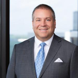 WLC alumnus in front of window with a city view in a gray suit with a blue tie.